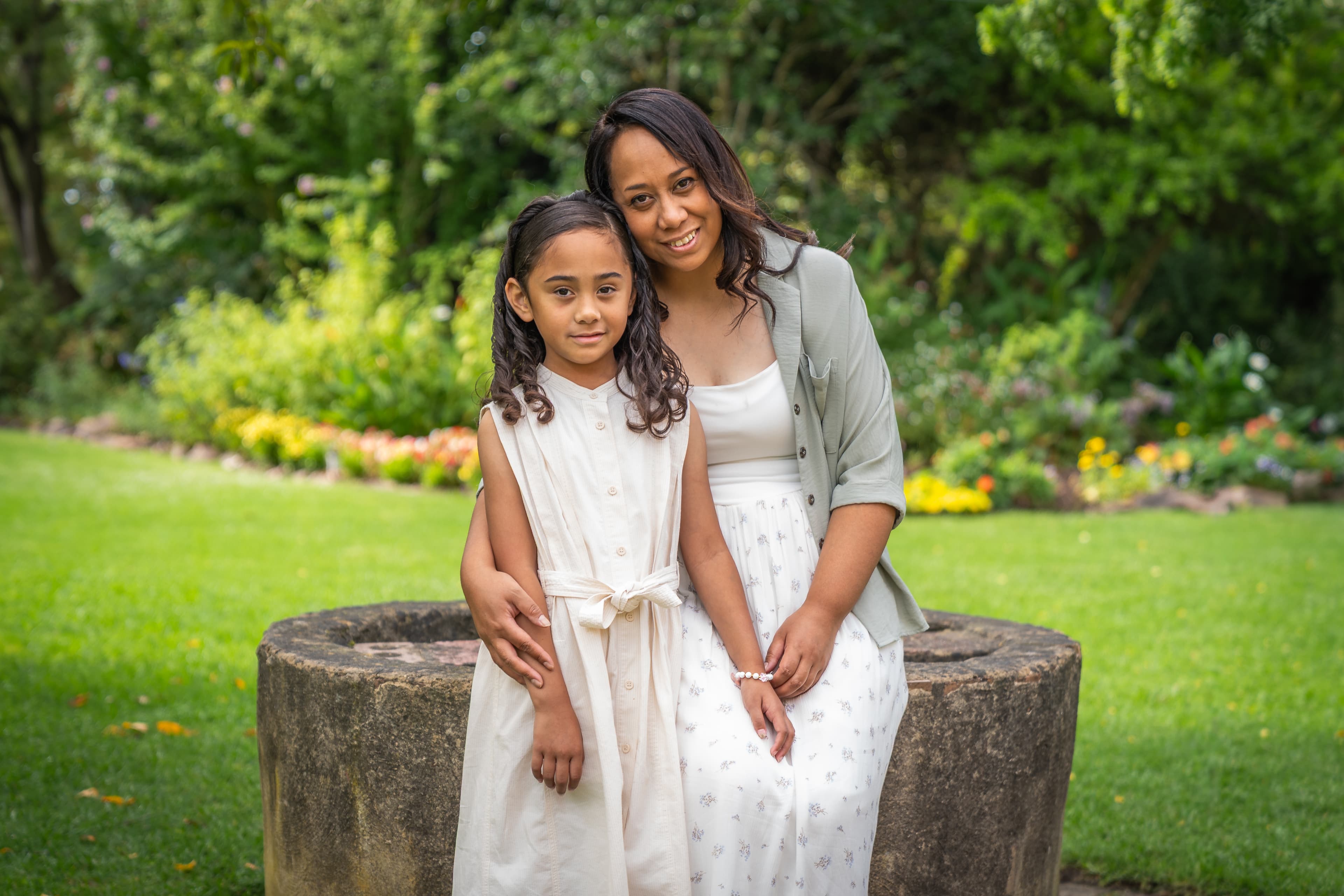 A smiling mother and daughter sit together on a stone well in a lush garden.